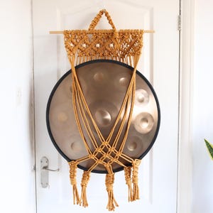 Pode incluir: Uma tapeçaria de macramé marrom dourado com um grande handpan redondo de cobre pendurado nela. O handpan é suspenso pelo macramé, que está preso a uma vareta de madeira.