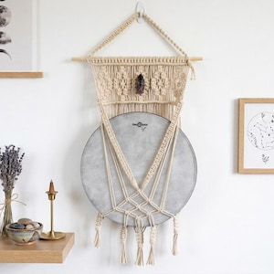 May include: A cream-colored macrame wall hanging featuring intricate knotted patterns and a central dark crystal, suspending a circular grey drum with the text "Remo" printed on the surface, finished with decorative fringe at the bottom edge.