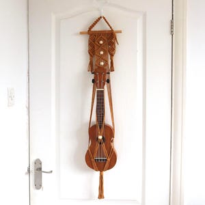 May include: A brown ukulele hangs from a macrame wall hanger on a white door. The macrame is tan and features decorative wooden beads. The ukulele is a light brown color and is suspended by the macrame.
