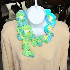 May include: A handmade crochet scarf in shades of green, blue, and white, displayed on a white mannequin. The scarf has a ruffled, spiral design, adding a unique texture. The background includes a beige shirt and dark wooden furniture.