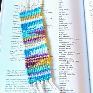 May include: A colorful woven bookmark with a fringe on the bottom. The bookmark is made of blue, green, purple, and yellow yarn. The bookmark is laying on a page of a cookbook with a list of ingredients.