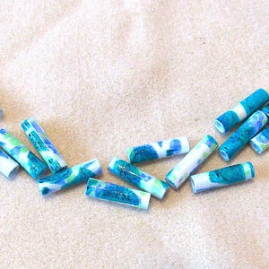 May include: A collection of cylindrical beads with a marbled design in shades of blue, teal, and white. The beads are scattered on a light beige surface, showcasing their colorful patterns and textures. These beads are suitable for jewelry making.