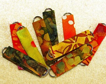 May include: A collection of rectangular fabric strips in various warm colors including red, orange, yellow, and green, each featuring batik-style patterns. Each strip is folded and secured with a silver-toned metal ring at both ends.