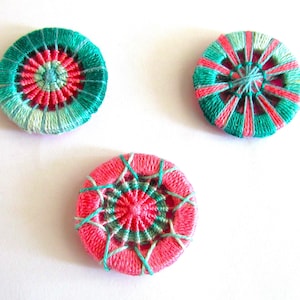 May include: Three handmade, colorful fabric buttons. Each button is round with a central design of pink and teal thread. The buttons are approximately 1.5 inches in diameter and are set against a white background.