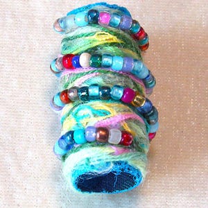 May include: A cylindrical object wrapped in multi-colored yarn in shades of green, yellow, and pink, featuring a spiral of small, translucent, and opaque glass beads in various colors including blue, red, purple, and metallic tones.
