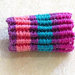 May include: A colorful, handmade scrunchie featuring a striped pattern. The scrunchie is made of a soft, textured material and displays bands of pink, teal, purple, and magenta. The accessory is designed to hold hair in a ponytail or bun.