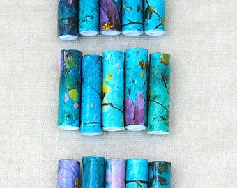 May include: Fifteen cylindrical paper beads arranged in three horizontal rows. Each bead features a vibrant blue base color with printed patterns of purple flowers, yellow accents, and dark brown branch-like lines across the surface.