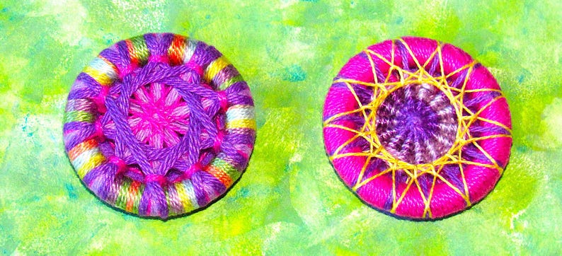 May include: Two colorful handmade buttons with a purple and pink center and a yellow and pink outer ring. The button on the left has a multi-colored outer ring, while the button on the right has a solid pink outer ring.