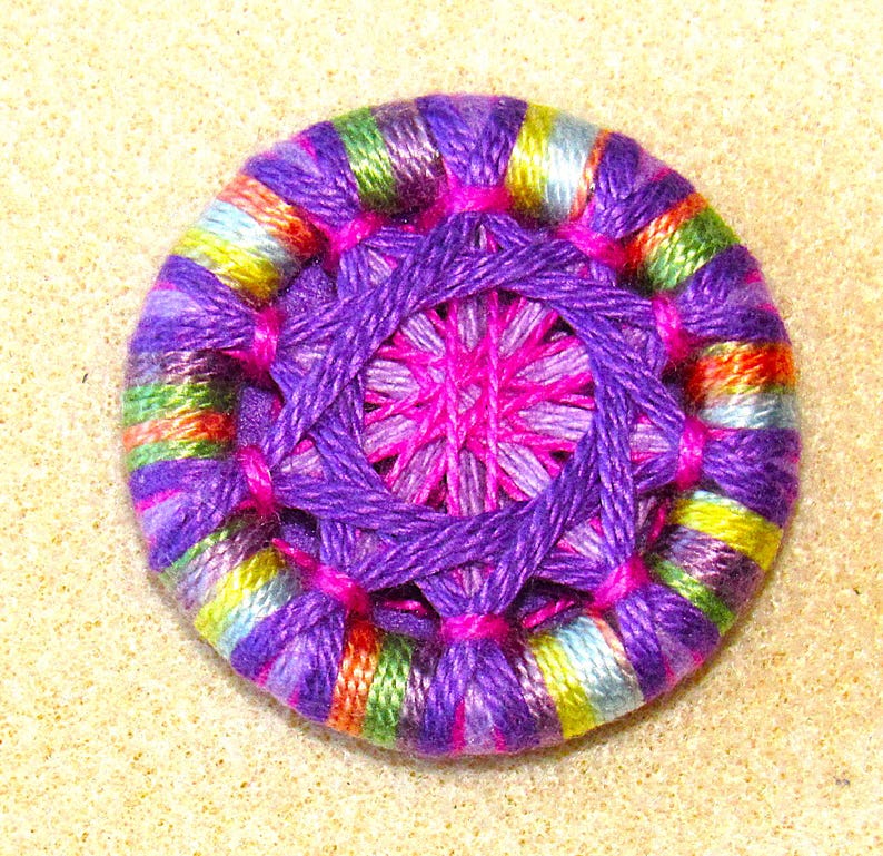 May include: A colorful, handmade button with a purple, pink, and green striped outer ring and a purple and pink center.