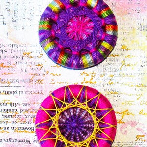 May include: Two colorful crocheted buttons with a purple center and a pink inner circle. The buttons are surrounded by a multi-colored yarn with yellow, green, blue, and pink accents.