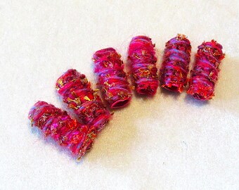 May include: Six cylindrical beads wrapped in vibrant magenta and hot pink yarn, featuring interwoven metallic gold fibers that create a textured, shimmering appearance. Each bead has a hollow center and a distinct, hand-wrapped fiber construction.