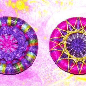 May include: Two colorful, round, embroidered buttons. One button is purple, pink, and green with a multi-colored center. The other button is pink with a purple center and gold accents.