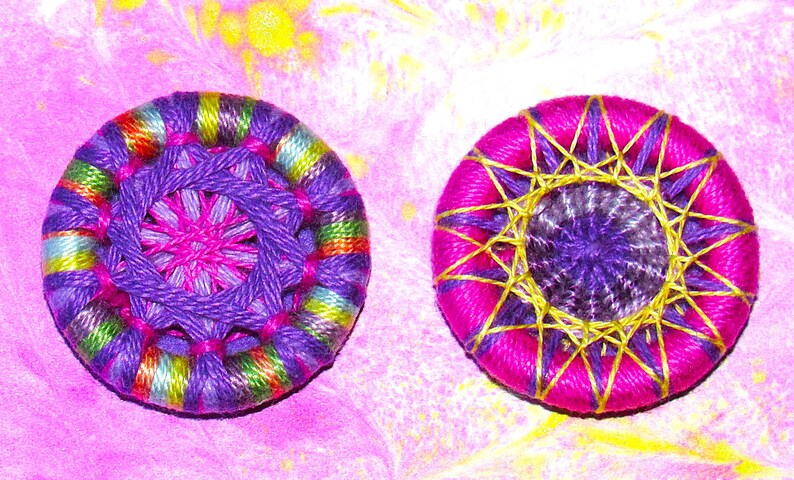 May include: Two colorful, round, embroidered buttons. One button is purple, pink, and green with a multi-colored center. The other button is pink with a purple center and gold accents.