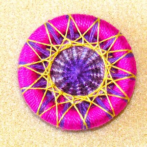 May include: A pink button with a purple and gold center. The button is decorated with a starburst pattern.