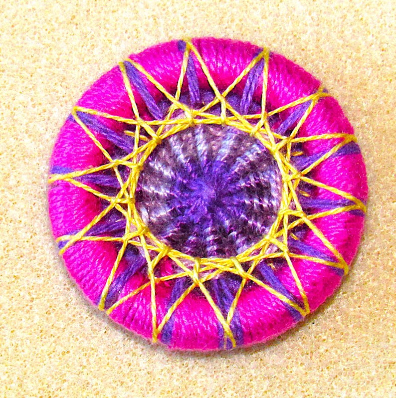 May include: A pink button with a purple and gold center. The button is decorated with a starburst pattern.