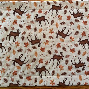 May include: A rectangular placemat with a repeating pattern of brown moose, autumn leaves, pine cones, and animal tracks on a cream background. The placemat is likely made of fabric and has a stitched border.