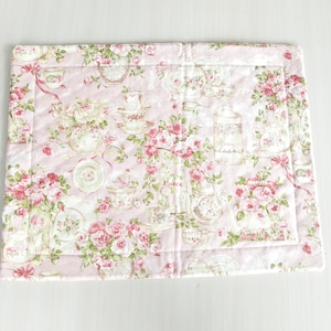 May include: A pink quilted placemat with a floral pattern featuring roses, teacups, and jars. The pattern is in shades of pink, green, and brown.