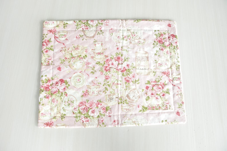 May include: A pink quilted placemat with a floral pattern featuring roses, teacups, and jars. The pattern is in shades of pink, green, and brown.