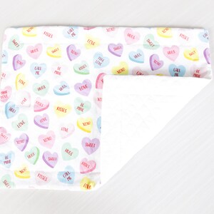 May include: A white cotton baby blanket with a colorful candy heart pattern. The hearts have various messages on them, including "love", "kisses", "hugs", "sweet", "call me", and "be mine".