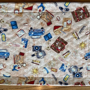 May include: A beige fabric placemat with a travel theme. The placemat features a repeating pattern of suitcases, cars, cameras, maps, and keys. The text "Road Trip" appears multiple times on the placemat.