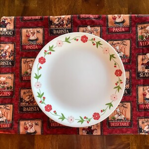 May include: A white ceramic dinner plate with a floral design border on a red and white quilted placemat with a repeating pattern of chefs and food-related words. The words on the placemat include "PASTA", "BARISTA", "VEGGIES", "SEAFOOD", and "DELECTABLE".