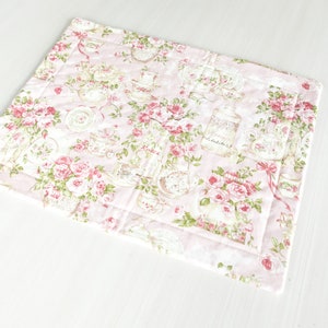 May include: A pink quilted placemat with a floral pattern featuring roses, teacups and jars. The placemat has a white border.