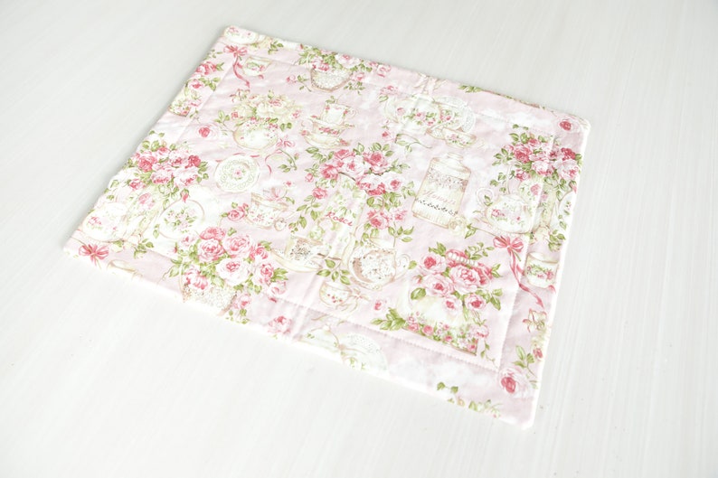 May include: A pink quilted placemat with a floral pattern featuring roses, teacups and jars. The placemat has a white border.