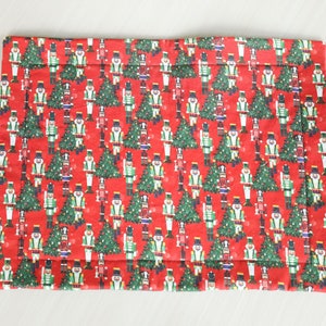 May include: A rectangular, red Christmas-themed placemat. The fabric is patterned with green Christmas trees and toy soldier nutcrackers in various poses. The placemat has a quilted appearance.