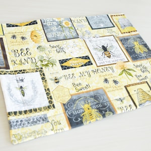 May include: A yellow and gray fabric placemat with a bee theme. The placemat features a variety of bee-related images and text, including bees, honeycombs, flowers, and the words "Bee Kind", "Save the Bees", and "Bee My Honey".