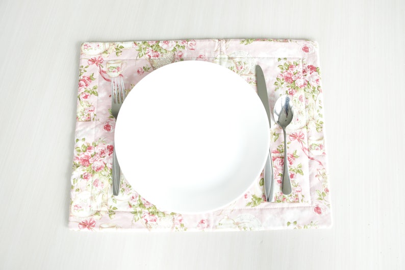 May include: A white ceramic plate with a silver fork, knife, and spoon on a pink floral placemat.