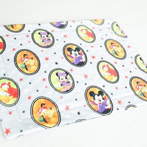 May include: A white fabric placemat with a repeating pattern of Mickey Mouse, Minnie Mouse, and Pluto dressed in Halloween costumes. The characters are in circles with a black outline and a light green background. The background of the placemat is white with red and black stars.