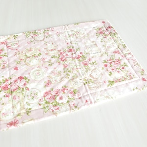 May include: A pink quilted placemat with a floral pattern featuring roses and teacups. The placemat has a white border and is made of fabric.