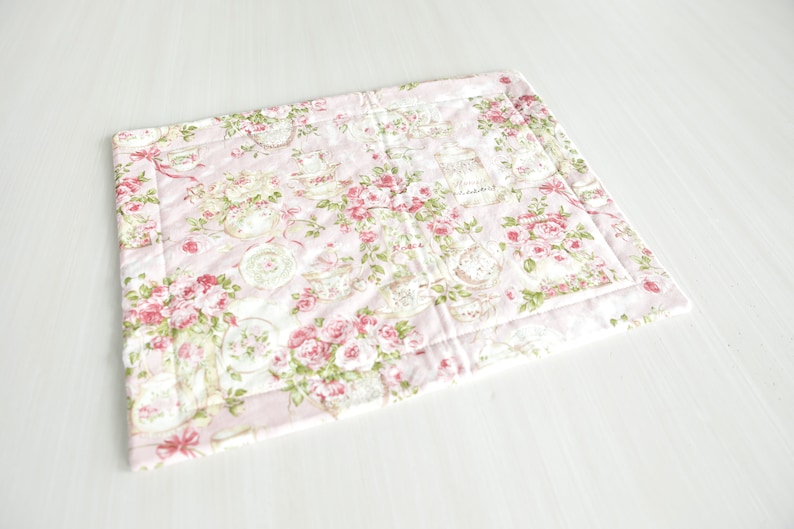 May include: A pink quilted placemat with a floral pattern featuring roses and teacups. The placemat has a white border and is made of fabric.