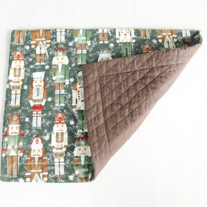 May include: A green quilted placemat with a pattern of nutcracker soldiers on a snowy background. The back of the placemat is brown and quilted.