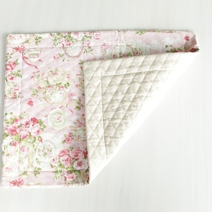 May include: A pink and white quilted placemat with a floral pattern. The placemat is folded in half, showing the quilted side.