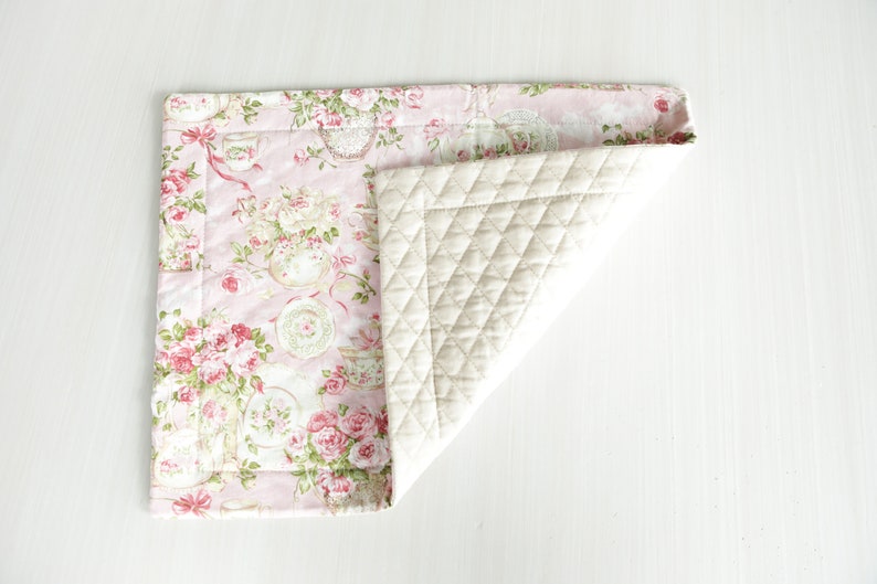 May include: A pink and white quilted placemat with a floral pattern. The placemat is folded in half, showing the quilted side.