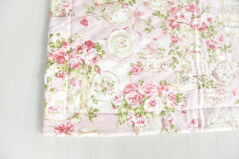 May include: Pink fabric with a floral pattern featuring roses, teacups, and teapots. The fabric has a quilted texture and a green border.