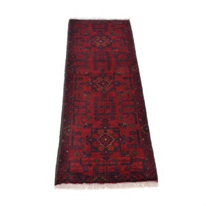 May include: A rectangular, red oriental rug with a detailed geometric pattern. The rug features a central design with repeating motifs in navy blue and touches of gold. The edges have a fringe.