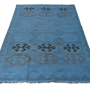 May include: A rectangular area rug in a vibrant blue hue featuring a repeating pattern of dark brown geometric shapes and stylised motifs. The rug has a flat-woven texture and is finished with short, straight fringe along the top and bottom edges.