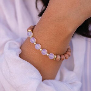May include: A beaded bracelet featuring light pink and brown beads with gold accents. The bracelet is worn on a wrist, with a white shirt and a blue and white striped top in the background.