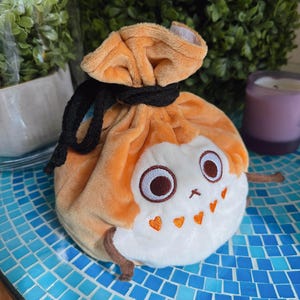 Puede incluir: Una bolsa de cordón de felpa naranja y blanca con una linda cara de dibujos animados y corazones. La bolsa tiene un cordón de ajuste negro y está hecha de tela suave y peluda.