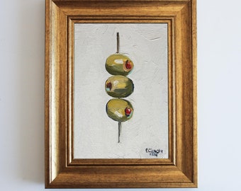 May include: An oil painting in a gold frame depicting three green olives with red pimentos skewered on a thin metal pick against a textured white background. The artwork features visible brushstrokes and a signature in the bottom right corner.