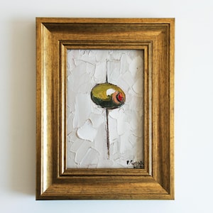 May include: An oil painting in a gold-toned frame featuring a green olive with a red pimento center, set against a textured white background with thick impasto brushstrokes. A thin vertical line extends from the olive, and a signature is visible.