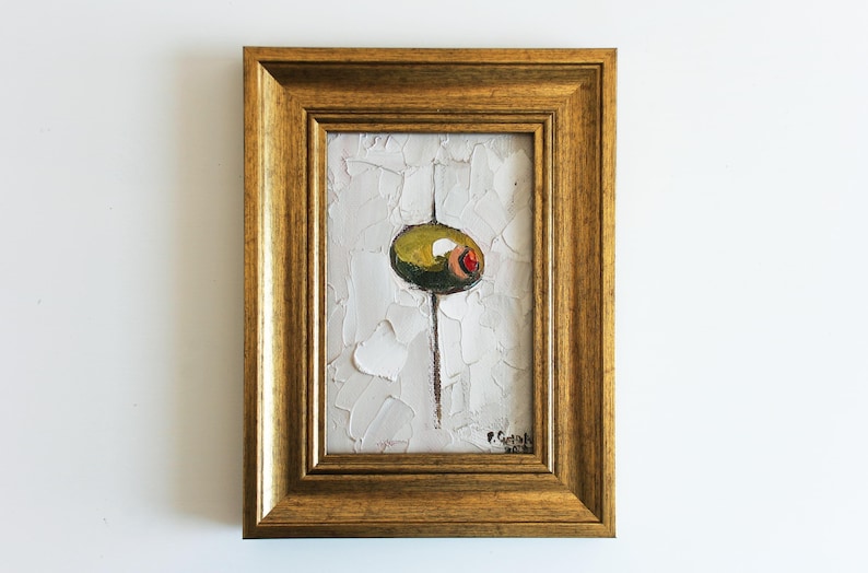 May include: An oil painting in a gold-toned frame featuring a green olive with a red pimento center, set against a textured white background with thick impasto brushstrokes. A thin vertical line extends from the olive, and a signature is visible.