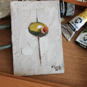 May include: A small rectangular oil painting on canvas featuring a single green olive with a red pimento center, centered on a thick, textured white impasto background. A thin brown vertical line extends downward from the olive. Signed in the corner.