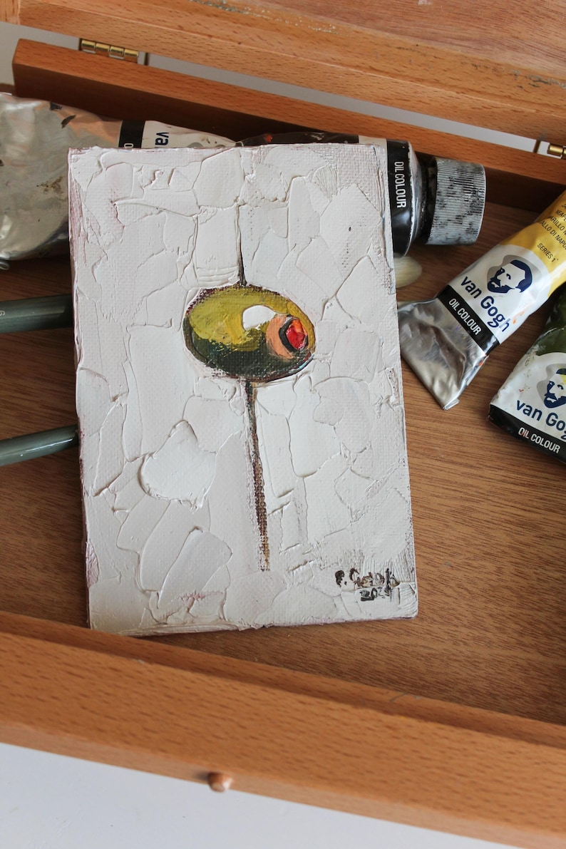 May include: A small rectangular oil painting on canvas featuring a single green olive with a red pimento center, centered on a thick, textured white impasto background. A thin brown vertical line extends downward from the olive. Signed in the corner.