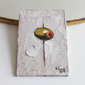 May include: A rectangular oil painting on canvas featuring a thick, textured impasto application of white paint. In the center, a single green olive with a red pimento is depicted, balanced on a thin vertical brown line. The artist's signature is visible.