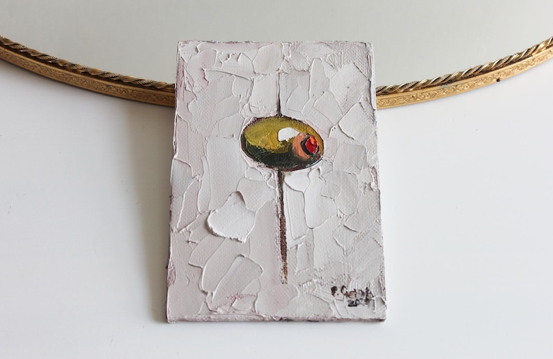 May include: A rectangular oil painting on canvas featuring a thick, textured impasto application of white paint. In the center, a single green olive with a red pimento is depicted, balanced on a thin vertical brown line. The artist's signature is visible.