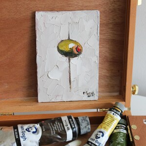 May include: A small rectangular oil painting on canvas featuring a single green olive with a red pimento center against a textured white background, displayed inside an open wooden artist box with tubes of Van Gogh oil paint visible below.