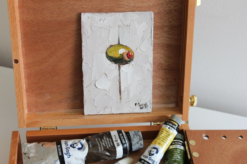 May include: A small rectangular oil painting on canvas featuring a single green olive with a red pimento center against a textured white background, displayed inside an open wooden artist box with tubes of Van Gogh oil paint visible below.